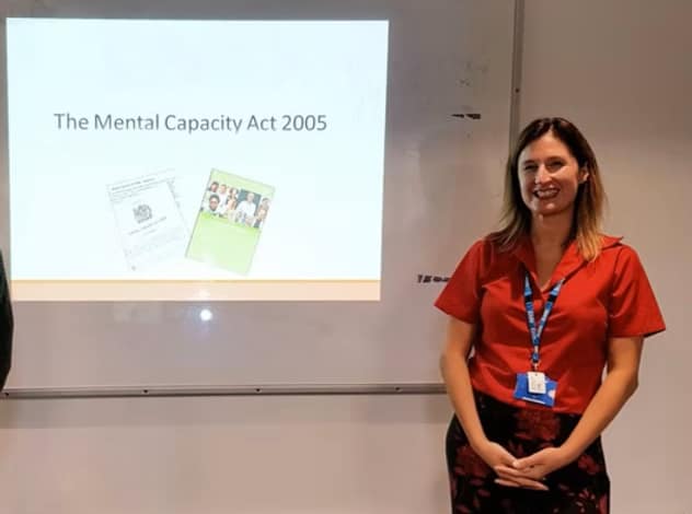 A picture of a women standing in front of a whiteboard which has a presentation projected onto it, the presentation shows the words "The Mental Capacity Act 2005".